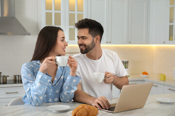 Canvas Print - Happy couple in pajamas with laptop having breakfast at kitchen table