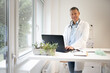 © epiximages - handsome doctor with white coat and blue shirt and stethoscope stands behind high table and works on laptop and has web conference or meeting