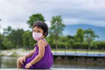 Wall Mural - Adorable Little girl wearing protective face mask sitting at lake outdoor.