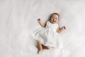 Little female baby lying on the bed and smiling. Girl dressed in beautiful white dress.