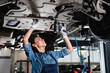 © LIGHTFIELD STUDIOS - young african american mechanic in overalls repairing bottom of car with electric screwdriver in garage