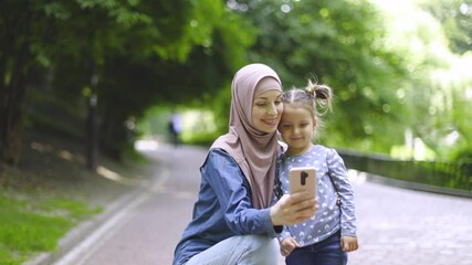 Wall Mural - Pretty happy young Muslim mother in hijab and cute little daughter, taking selfie photo with phone camera, or making video call to friend, outdoor in the summer park on sunny day