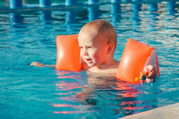 Cute little baby boy playing in outdoor swimming pool on hot summer day. Kids learn to swim. Happy child with orange floaties. Swimming aid protection for kid. Family summer vacation.