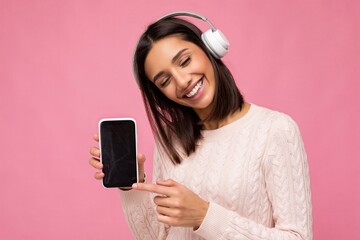 Wall Mural - Beautiful cute young brunette woman wearing pink sweater isolated over pink background wall wearing white bluetooth wireless headphones and listening to good music and showing mobile phone with empty