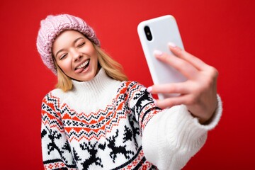 Photo of pretty positive young blonde woman wearing warm knitted hat and winter warm sweater standing isolated over red background using mobile phone taking selfie photo looking at device display and