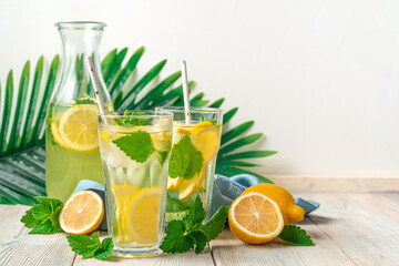 Refreshing lemonade in two glasses and a decanter, lemons and mint on a white background with palm branches.