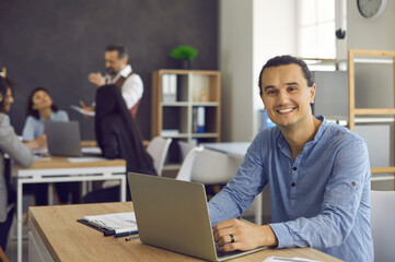 Wall Mural - Portrait of confident young business owner. Successful millennial man sitting at desk with laptop computer looking at camera with genuine happy smile. Coworking office workspace copy space background