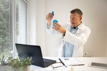 male chemist, scientist works at standing table and examines blue liquids in small vials and wears white coat