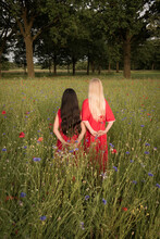 Two Girl Teenagers, Blonde And Black Long Hair, Wearing Red Dresses In Field Of Wild Poppy Flowers Seen From Back