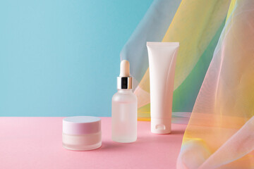 serum, facial cleanser and moisturizer cream on blue pink background with colorful rainbow organza f