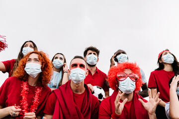 Multiracial hispanic football supporters wearing protective face mask watching soccer match event at stadium - New normal lifestyle in sport events - Copy space 