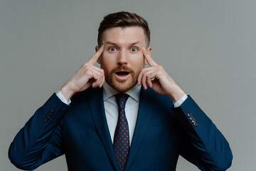 Wall Mural - Amazed businessman in suit holding fingers on temples and looking at camera with shocked face expression