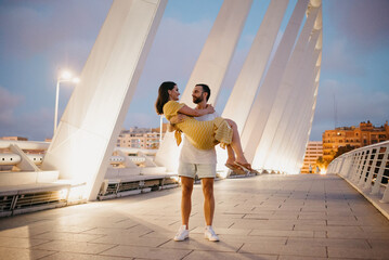 Wall Mural - A man with a beard is holding his girl in a yellow dress in his arms on a white bridge in Valencia. A couple of tourists in full length on a date in the warm evening.