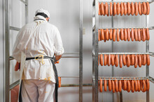 Worker Hangs Raw Sausages On Racks In Storage Room At Meat Processing Factory. High Quality Photo.