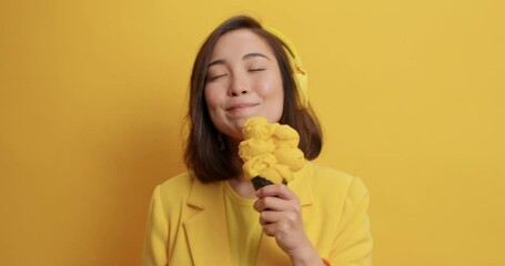 Wall Mural - Pretty beautiful Asian woman with dreamy expression has pleasant thoughts eats delicious ice cream during summer time listens audio track via headphones isolated over vivid yellow background