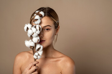 Portrait of pretty young woman with branch of soft cotton with clean fresh skin in studio against beige background. Cosmetology, beauty and spa concept.