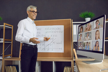 Happy male teacher having online class with group of distant students. Smiling mature man standing in front of classroom board and giving virtual math lesson via video conference on desktop computer