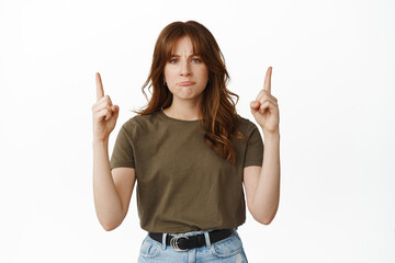 Wall Mural - Unfair. Frowning upset girl complains, pointing fingers up and sulking, showing disappointing news, express regret or being jealous, standing against white background