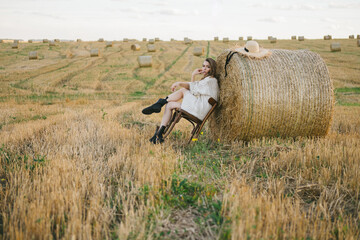 beautiful woman in a jacket and a hat with a chair by a haystack in the field.