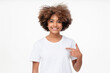 © Damir Khabirov - African student girl pointing with index finger at blank white t-shirt with empty space for your advertising text or image, standing isolated on studio background