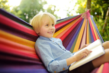 cute little blond caucasian boy reading book and having fun with multicolored hammock in backyard or