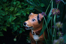 Cute Ceramic Dog Figurine. Spring Garden After Rain With Garden Statue. Dark Natural Background, Soft Focus