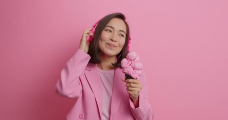 Wall Mural - Happy Asian woman licks ice cream listens music via headphones eats delicious summer dessert has carefree mood dressed in fashionable clothes isolated over pink background. What tasty gelato