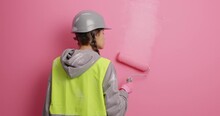 Room Interior Update. Busy Faceless Female Decorator Paints Wall In Pink Dye With Roller Stands Back Then Turns At Camera Has Annoyed Irritated Expression Uses Repair Tools Wears Helmet And Uniform