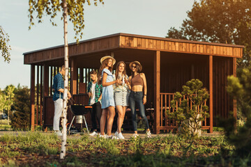 Wall Mural - Young friends grilling meat while enjoying barbecue, group of happy guys and girls cooking and eating on barbecue dinner outdoors