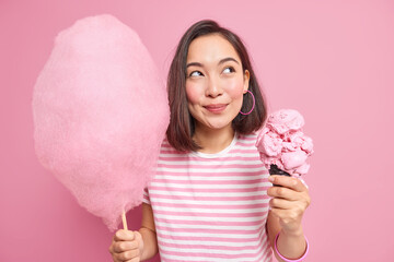 Wall Mural - Studio shot of good looking Asian woman with dark hair has dreamy expression holds sweet cotton candy and ice cream thinks about something pleasant poses against pink background dressed casually