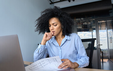 Wall Mural - Concerned African American businesswoman ceo analyst sitting at desk reading papers financial reports with laptop in contemporary corporation office. Financial data analyst working with big data.