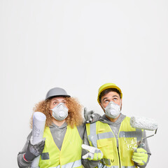 Two maintenance workers in safety clothes concentrates above notice something strange on ceiling. Woman architect and man painter with roller pose against white background at construction site