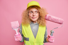 Repair Service. Pleased Thoughtful Woman Painter With Curly Fair Hair Holds Brush And Roller Thinks About Interior Design Of New Dwelling Does Manual Work Dressed In Uniform Protective Helmet