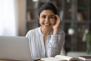 Wall Mural - Office worker. Positive young mixed race woman employee manager pose for portrait by work desk with pc paper notebook. Teenage indian female student look at camera with smile distracted from paperwork