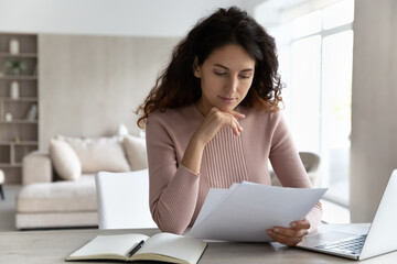 Poster - Pensive young Hispanic woman sit at desk at home office work online on computer read paperwork documents. Millennial Latino female use laptop consider financial report or paper correspondence.