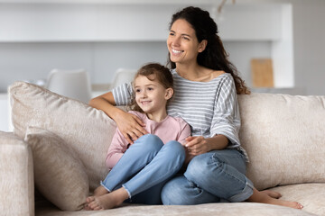 Wall Mural - Smiling Hispanic young mother and happy small biracial daughter sit on sofa at home look in distance dream visualize together. Excited Latino mom and little ethnic girl child relax. Vision concept.