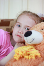 Little Girl With Sad Eyes Looks At The Camera Sits With An Orange Bear Toy