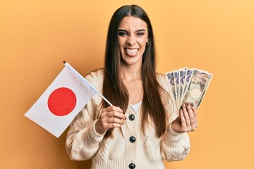 Wall Mural - Beautiful brunette young woman holding japan flag and yen banknotes sticking tongue out happy with funny expression.