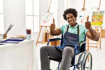 Young african american artist man sitting on wheelchair at art studio approving doing positive gesture with hand, thumbs up smiling and happy for success. winner gesture.