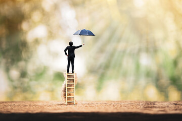 Businessman stand on the gold coin with growing interest and hand hold open the black umbrella for protect of success in the public park, to prevent for money and business investment of asset concept.