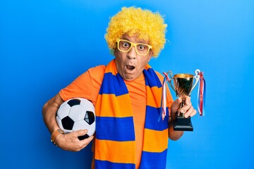 Poster - Senior hispanic man football hooligan cheering game holding ball and trophy in shock face, looking skeptical and sarcastic, surprised with open mouth