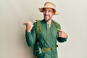 Poster - Handsome man with beard wearing explorer hat and backpack smiling with happy face looking and pointing to the side with thumb up.