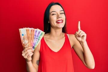 Poster - Young hispanic girl holding philippine peso banknotes smiling with an idea or question pointing finger with happy face, number one