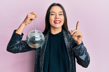 Poster - Young hispanic girl holding shiny disco ball smiling with an idea or question pointing finger with happy face, number one