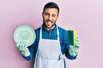 Poster - Young hispanic man wearing apron holding scourer washing dishes winking looking at the camera with sexy expression, cheerful and happy face.