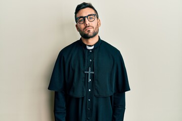 Canvas Print - Young hispanic man wearing priest uniform standing over white background relaxed with serious expression on face. simple and natural looking at the camera.