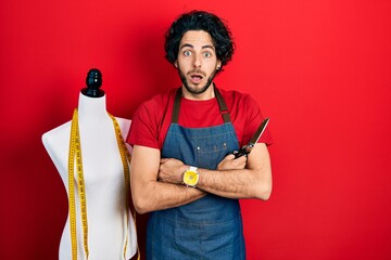 Poster - Handsome hispanic man standing by manikin holding scissors afraid and shocked with surprise and amazed expression, fear and excited face.