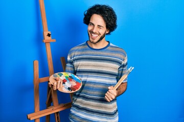 Poster - Handsome hispanic man standing drawing with palette by painter easel stand winking looking at the camera with sexy expression, cheerful and happy face.