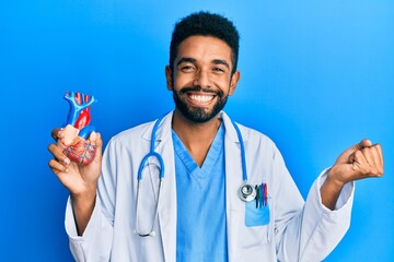 Wall Mural - Handsome hispanic man with beard wearing doctor uniform holding heart screaming proud, celebrating victory and success very excited with raised arm