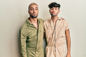 Poster - Homosexual gay couple standing together wearing casual jumpsuit looking at the camera blowing a kiss on air being lovely and sexy. love expression.